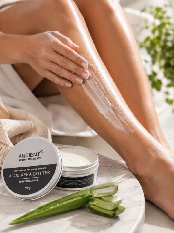 Woman applying Aloe Vera & Coconut body butter to her leg beside an open tin and fresh aloe leaves.