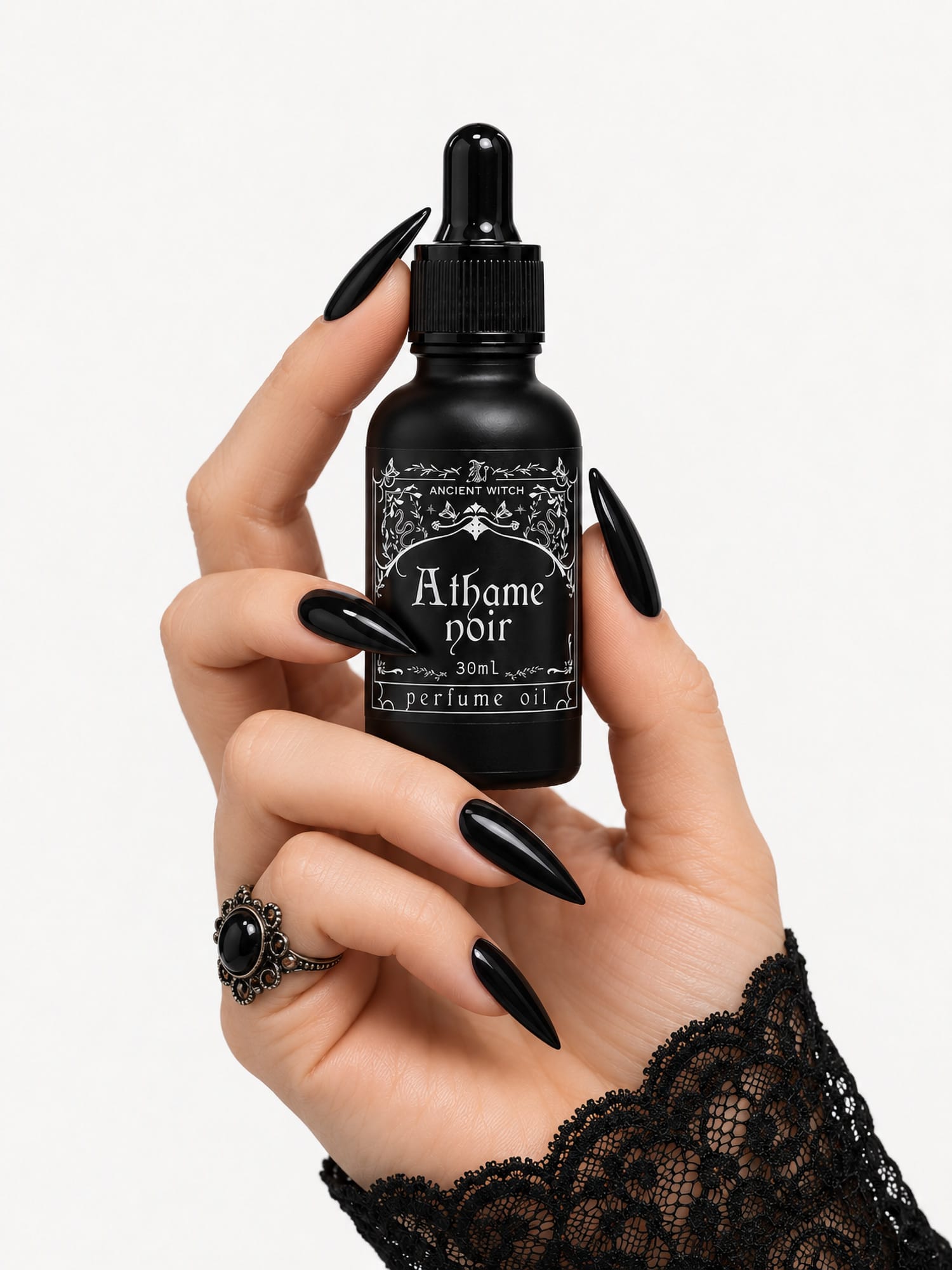 Hand with black pointed nails holding a 30ml bottle of Ancient Witch Athame Noir perfume oil against a white background.